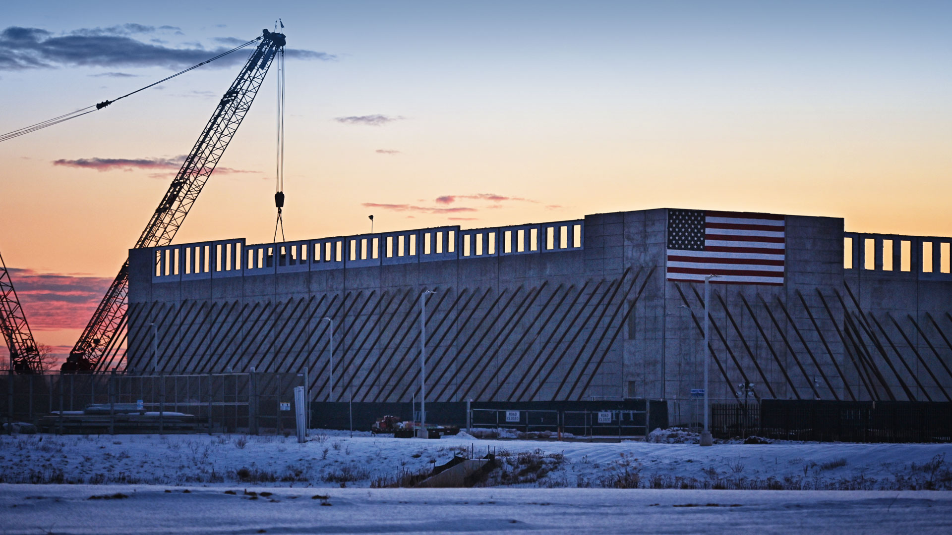 Large concrete structure under construction at sunset, with two cranes positioned nearby and an American flag displayed on the building’s wall. Snow covers the ground in the foreground.
